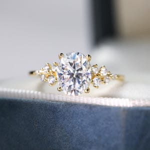 Puede incluir: Un anillo de compromiso de oro con una piedra central de forma ovalada y piedras más pequeñas engastadas en un delicado diseño floral.