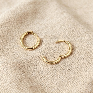 May include: A pair of gold hoop earrings with a smooth, polished finish. One earring is a complete circular hoop, while the other is a hinged hoop earring shown in an open position, revealing the thin post mechanism used for closure.