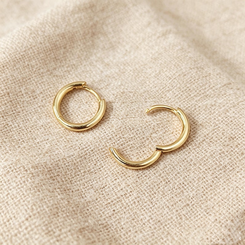 May include: A pair of gold hoop earrings with a smooth, polished finish. One earring is a complete circular hoop, while the other is a hinged hoop earring shown in an open position, revealing the thin post mechanism used for closure.