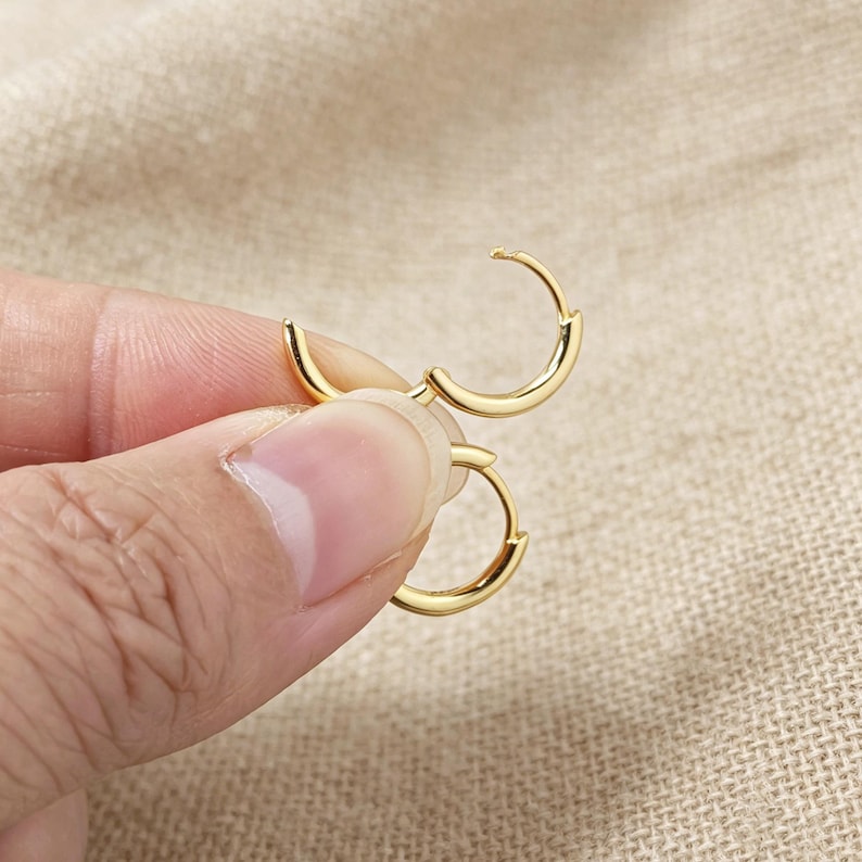May include: A pair of small, polished gold-tone hoop earrings. Each earring features a thin, circular metal band with a smooth, reflective surface and a simple hinged closure mechanism, designed for a secure fit in a pierced earlobe.