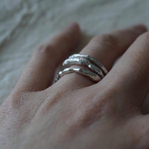 May include: Three silver rings with a textured, organic design are worn on a finger. The rings have a slightly irregular shape, giving them a handcrafted appearance. The rings are stacked together, creating a layered effect.
