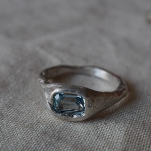 May include: A silver ring with a light blue rectangular gemstone. The ring has a textured, organic design. The gemstone is set in a bezel setting, and the band has a slightly uneven surface.