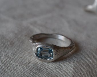 May include: A silver ring with a light blue rectangular gemstone. The ring has a textured, organic design. The gemstone is set in a bezel setting, and the band has a slightly uneven surface.