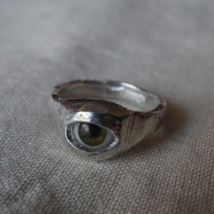 May include: A silver ring with a textured band and a detailed eye design. The eye features a green iris and a white sclera, creating a unique and artistic piece of jewelry. The ring's design is eye-catching and unusual.