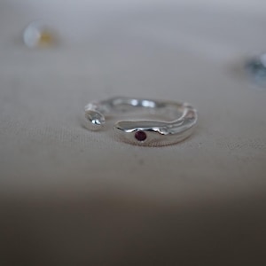 Peut inclure: Bague argentée avec un design ouvert. La bague a une surface texturée et une petite pierre précieuse rouge ronde sertie dans le groupe. La bague est présentée sur une surface de couleur neutre.