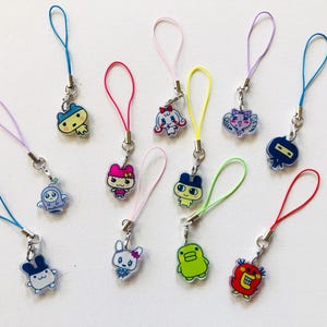 May include: A collection of eleven small acrylic charms featuring various colorful cartoon characters, each attached to a thin, vibrant nylon cord loop in shades of blue, pink, yellow, purple, red, and green, designed for attachment to mobile devices.