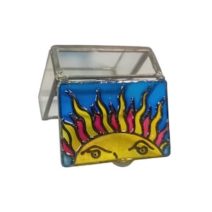 Könnte beinhalten: Kleine, aufklappbare Glasbox mit farbenfrohem Sonnendesign. Die Sonne hat ein gelbes Gesicht vor blauem Hintergrund mit gelben, rosa und roten Flammen. Die Box besteht aus klarem Glas mit Metallkanten.