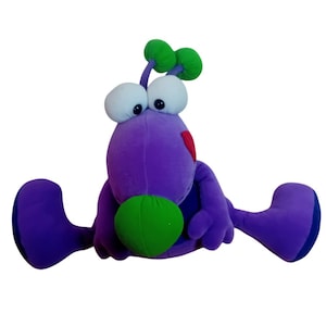 Puede incluir: Un juguete de peluche morado con ojos grandes, una antena verde y un corazón rojo en el pecho. El juguete tiene dos brazos verdes y dos piernas moradas.