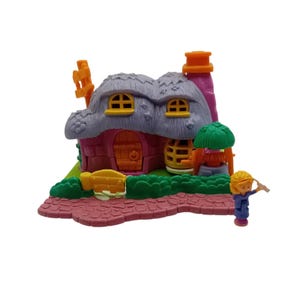 Può includere: Un cottage in miniatura rosa e grigio con tetto di paglia, finestre gialle e un camino. Una piccola figura in blu e giallo è nelle vicinanze. Il cottage ha una porta di legno e un piccolo pozzo.