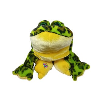 Puede incluir: Un juguete de peluche con forma de rana, con una combinación de colores verde y amarillo. La rana tiene ojos grandes y redondos y una expresión sonriente. El cuerpo es una mezcla de manchas verdes y marrones, con detalles amarillos en el vientre y los pies. Un logotipo colorido está en el pie de la rana.
