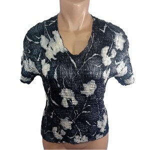 May include: A short-sleeved, V-neck top with a textured, pleated fabric. The top features a dark navy blue background with a white floral pattern. The fabric appears to have a crinkled texture, adding visual interest to the garment.