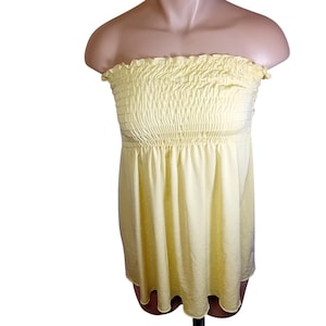 May include: A pale yellow strapless tube top with a smocked bodice and a flowing, slightly textured fabric. The top has a gathered neckline and a loose, flared bottom, creating a casual and summery look. The fabric appears to be lightweight.