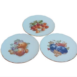 May include: Three white ceramic plates with gold trim, each featuring a different fruit design. The first plate has strawberries, the second has peaches and plums, and the third has plums and grapes.
