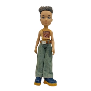 Könnte beinhalten: Eine Plastikpuppe mit braunen Haaren, die ein gelbes Tanktop mit einem grafischen Aufdruck, eine blaue Jeans und blaue und gelbe Schuhe trägt.