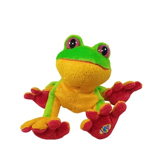 May include: A plush frog toy with a green head, yellow body, and red feet. The frog has large, pink eyes and is posed with its arms and legs outstretched. The toy has a sparkly texture and a small logo on one foot.