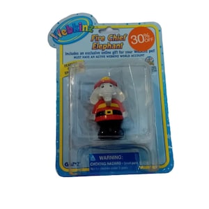 Puede incluir: Un juguete Webkinz Fire Chief Elephant en un paquete de plástico. El elefante lleva un uniforme rojo de jefe de bomberos con un cinturón negro y un casco amarillo. El paquete tiene una pegatina de venta con un 30% de descuento.
