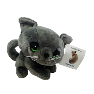 May include: A plush gray cat toy with large green eyes and white whiskers. The toy has a tag that reads "Russian Blue" and features a small illustration of a cat. The tag also includes the text "THE CAT".