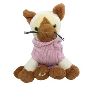 May include: A soft toy Siamese cat with brown markings and a pink knitted sweater. The cat has green eyes and black whiskers. The toy is sitting upright with its paws forward.