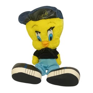 May include: A plush Tweety Bird toy wearing a denim cap, black shirt, and blue trousers. The yellow bird has blue eyes and an orange beak. The shoes are black with white stitching and soles. The cap has a black leather patch with a Tweety Bird graphic.
