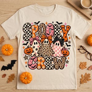 Op de afbeelding: Een beige t-shirt met korte mouwen en een grafische print met de tekst SPOOKY CREW. Het ontwerp bevat drie spoken met strikken, omringd door pompoenen, vleermuizen, sterren en patronen met luipaardprint en zwart-wit geblokte motieven.