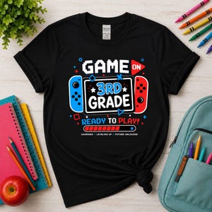 May include: A black short-sleeve t-shirt featuring a graphic of a video game console with the text GAME ON 3RD GRADE READY TO PLAY. The design includes blue and red controller sides, a loading bar, and the words LEARNING LEVELING UP FUTURE UNLOCKED.