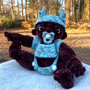 May include: A crocheted plush toy with a dark purple body, blue and white crocheted clothing, and a pacifier. The toy has horns and a blue crocheted hat.