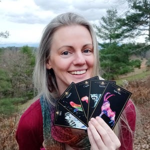 May include: A woman holding a fan of tarot cards with colorful illustrations. The cards feature images of animals and celestial designs. The background includes trees and a cloudy sky.