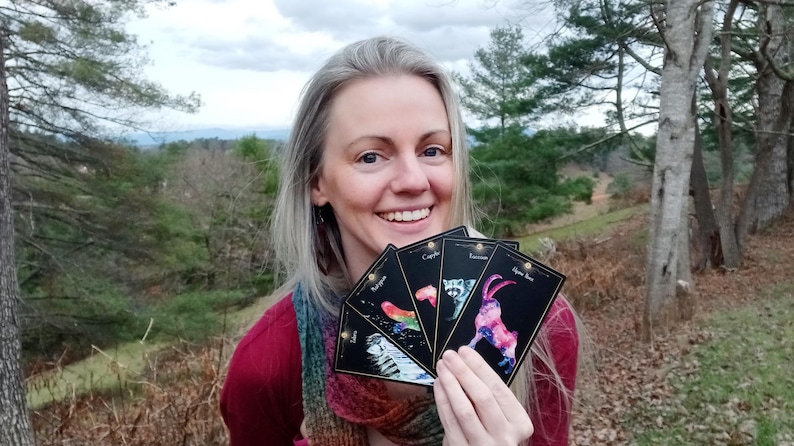 May include: A woman holding a fan of tarot cards with colorful illustrations. The cards feature images of animals and celestial designs. The background includes trees and a cloudy sky.