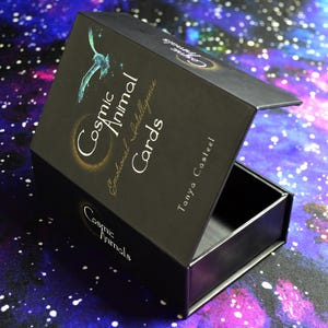 May include: Black box with the text "Cosmic Animal Cards" and a teal bird illustration. The box is open, revealing an empty interior. The background is a colorful galaxy design.