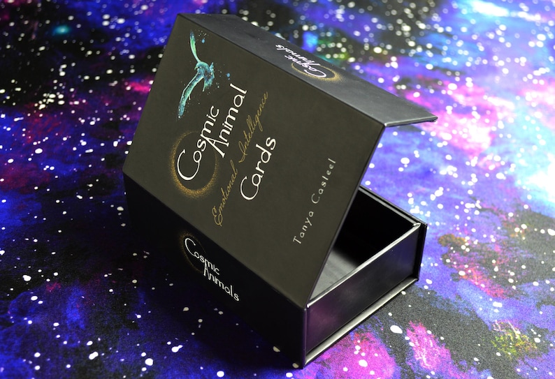 May include: Black box with the text "Cosmic Animal Cards" and a teal bird illustration. The box is open, revealing an empty interior. The background is a colorful galaxy design.