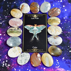 May include: A collection of oval-shaped stones surrounds a black card with a Luna Moth illustration and the word "Order." The stones vary in color, including beige, brown, gray, and pink. The background is a purple and blue galaxy design.