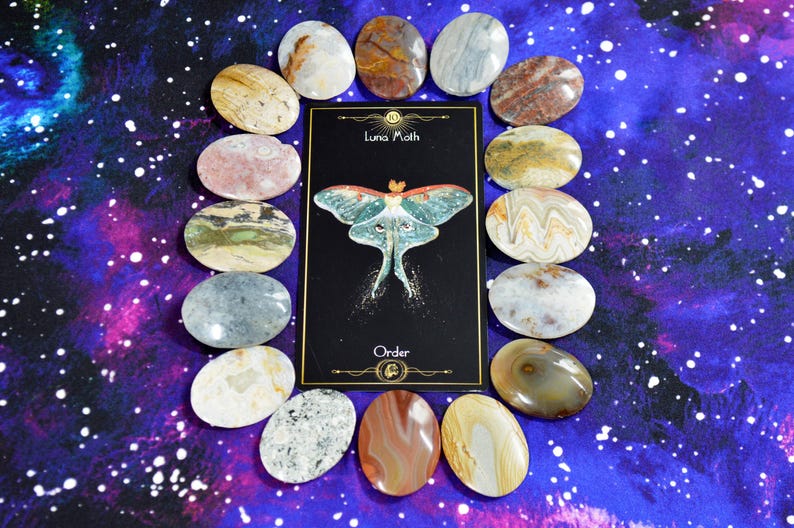 May include: A collection of oval-shaped stones surrounds a black card with a Luna Moth illustration and the word "Order." The stones vary in color, including beige, brown, gray, and pink. The background is a purple and blue galaxy design.