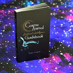 May include: A black guidebook titled "Cosmic Animal Emotional Intelligence Guidebook" with a teal bird illustration. The book is set against a backdrop of a starry, galaxy-themed surface.