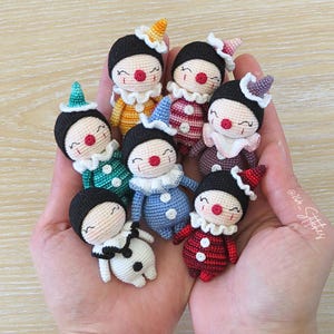 May include: A collection of seven crocheted clown dolls in various colors, including red, blue, green, yellow, and purple. Each doll has a black yarn hair, a red nose, and a white yarn collar.
