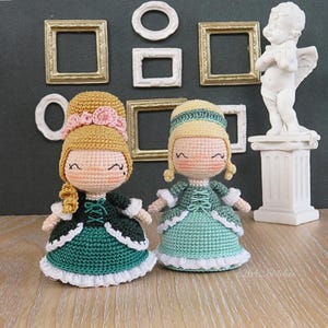 May include: Two handmade crochet dolls in teal and green dresses with white trim. The dolls have blonde hair and are smiling. The background includes small gold frames, white frames, and a white statue of an angel.