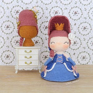 May include: Crocheted doll and hat set. The doll wears a blue dress with pink accents and a gold crown. The hat is pink and gold, with butterfly embellishments. The doll is approximately 15 cm tall.