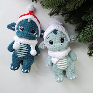 May include: Two crocheted dragon plushies. One is teal with a red and white Santa hat and the other is light green with a white hat. Both have white scarves and are decorated with pink accents. The plushies are next to a green pine branch.