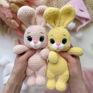 Peut inclure: Deux lapins en crochet faits à la main, un rose clair et un jaune, sont présentés sur un fond doux. Les lapins ont de grands yeux, des nez roses et de longues oreilles. Un chapeau rose en tricot avec des pompons blancs est en arrière-plan.