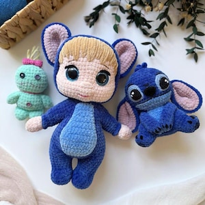 Pode incluir: Três bonecas de crochê feitas à mão. A boneca central é azul com uma barriga azul clara e olhos grandes. À esquerda, há uma boneca verde menor com um laço rosa. À direita, há uma boneca azul com orelhas grandes.