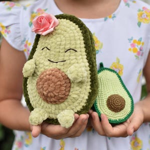 Puede incluir: Dos peluches de aguacate de crochet. Un aguacate es verde con un hueso marrón y una flor rosa en la parte superior. El otro aguacate es verde con un hueso marrón. Ambos aguacates tienen caras sonrientes.