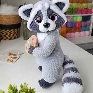 May include: A plush raccoon toy made of soft grey and white yarn, featuring large pink eyes, a black nose, and a striped tail. The raccoon is holding a small, round, tan-colored cookie decorated with colorful sprinkles in its dark grey paws.