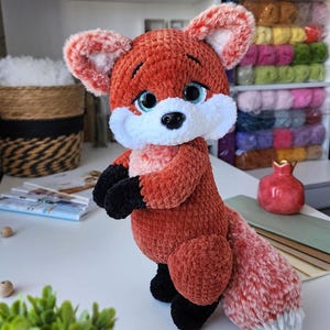 May include: A crocheted fox toy in shades of orange, white, and black. The fox features large blue eyes, a white muzzle, and a fluffy tail. The toy is made from soft yarn and stands on its hind legs.