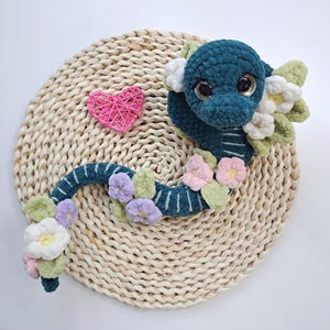 Peut inclure: Un serpent en crochet fait à la main, de couleur sarcelle, avec de grands yeux expressifs et des rayures blanches. Le serpent est orné de fleurs roses, violettes et blanches et de feuilles vertes. Un cœur rose tissé est également présent.