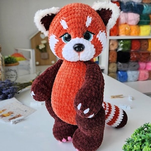 May include: A handmade crochet red panda plush toy. The toy has an orange body, dark brown limbs, and a striped tail. It features white accents on the face and ears, and blue eyes. The plush is standing on a white surface.