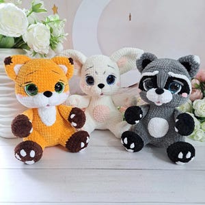 May include: Three crocheted soft toys: a fox, a bunny, and a raccoon. The fox is orange with brown details, the bunny is white with pink, and the raccoon is grey with black. Each has large, expressive eyes.
