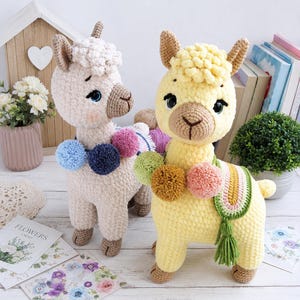 Puede incluir: Dos muñecos de llama tejidos a ganchillo hechos a mano con cuerpos de hilo texturizado. La llama de la izquierda es de color crema con pompones azules, azul marino y rosas. La llama de la derecha es amarillo pálido con pompones verdes, marrones y rosas.