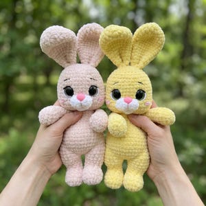 May include: Two plush crochet bunnies, one in soft pastel pink and the other in bright yellow. Each stuffed animal features long upright ears, large dark safety eyes, a small pink nose, and a white muzzle, crafted from textured yarn.