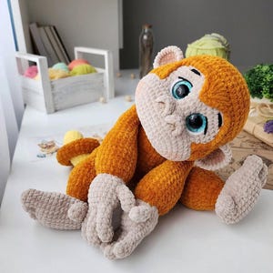 May include: A handmade crochet monkey doll with orange fur and light tan face, hands, and feet. The monkey has large blue eyes and is posed sitting down. The doll is a soft, plush toy.