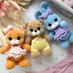 Könnte beinhalten: Drei handgefertigte Häkel-Plüschtiere, bestehend aus einer orangefarbenen Katze mit rosa Rüschenkleid und Schleife, einem braunen Bären mit gelber Latzhose und einem blauen Hasen mit lila Rüschenkleid und Schleife. Jedes Tier hat große, dunkle Augen.