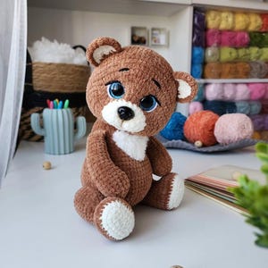 Puede incluir: Un oso de peluche marrón hecho a mano con detalles blancos en el hocico, el pecho y las almohadillas de los pies. El oso tiene grandes ojos azules y una nariz negra. El juguete de peluche está sobre una superficie blanca, mostrando su aspecto suave y texturizado.